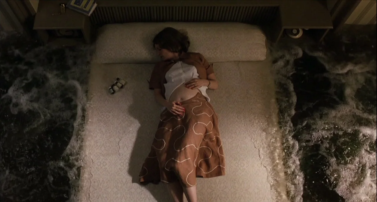 The Hours (2002) movie still: fullbody — A top-down view of a woman lying on a bed in a flooded room. Dark, turbulent ocean water surges on both…