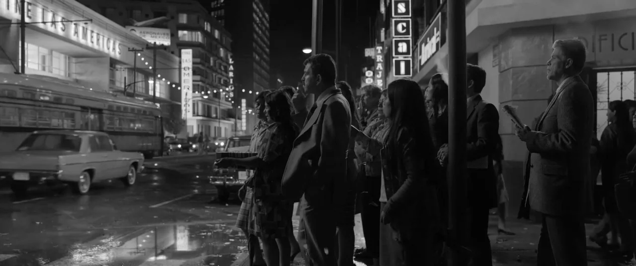 Roma (2018) movie still: wide — A busy 1970s urban intersection at night features neon-lit storefronts and movie theater marquees. A group of people…