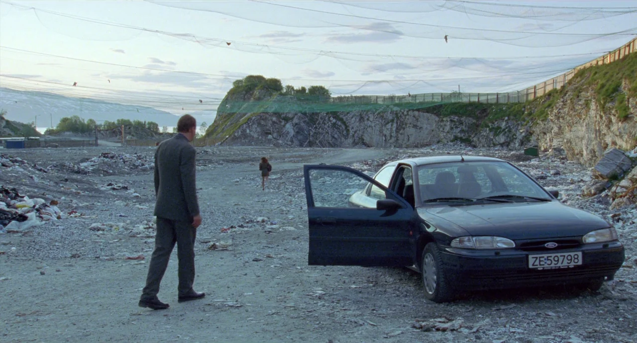 Insomnia (1997) movie still: wide — An expansive gravel quarry is enclosed by a high perimeter fence and netting. A man walks across the uneven…