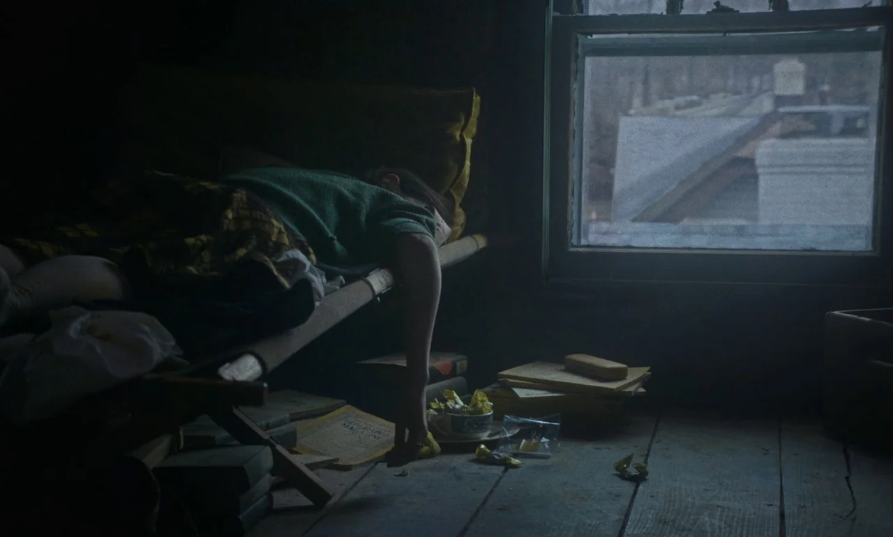 Eileen (2023) movie still: medium — A dark, cramped attic features a wooden cot positioned beside a small frosted window. A child wearing a green…