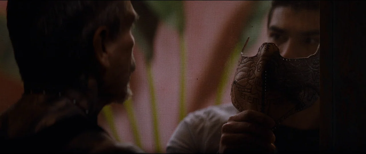 El Chicano (2018) movie still: medium — A man looks through a wire mesh screen while holding a carved wooden mask. The mesh creates a fine grid…
