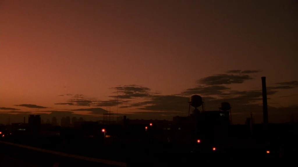 Cronos (1993) movie still: establishing — An industrial skyline appears in silhouette against a deep orange, dusk-lit horizon. Tall chimneys and…
