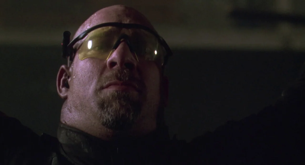 Universal Soldier: The Return (1999) movie still: closeup — A man in a dark environment looks down towards the camera lens. He wears yellow-tinted…