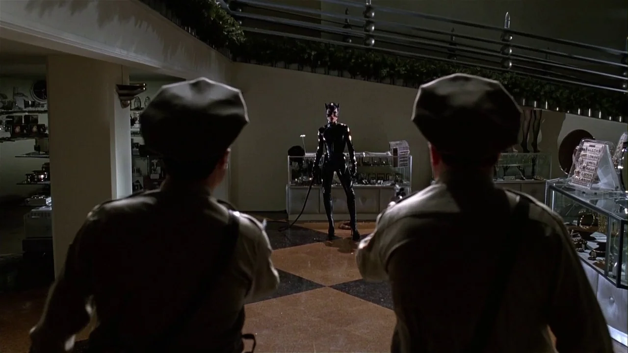 Batman Returns (1992) movie still: medium — A luxury boutique interior features glass display cases and a staircase with metal railings. A woman in…