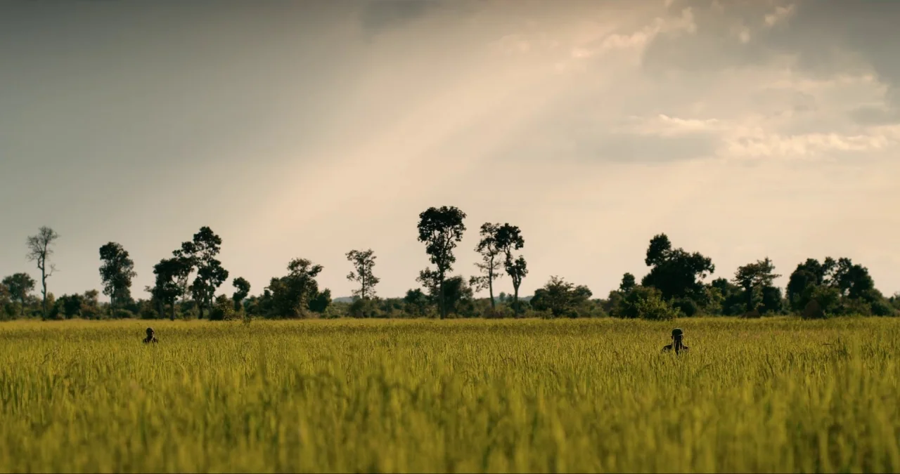 First They Killed My Father (2017) movie still: wide — A vast, flat grassy field stretches toward a distant, sparse line of dark trees. Two small…