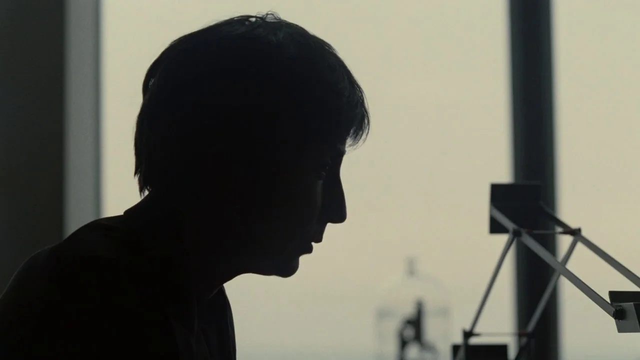 Splice (2009) movie still: closeup — A high-rise room features a large window with a bright, overcast sky outside. A person in profile sits in…