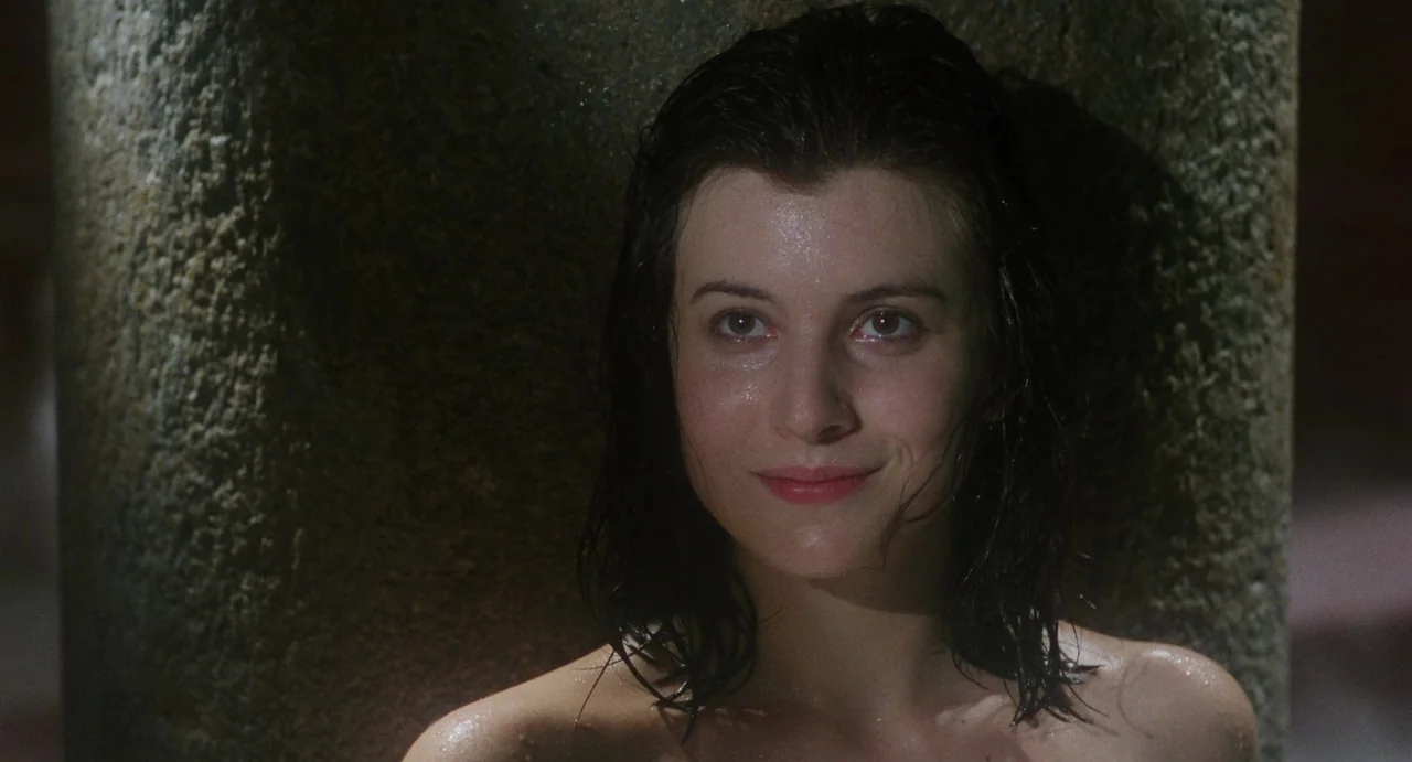The Spider Labyrinth (1988) movie still: closeup — A woman stands beside a large, dark stone pillar in a dimly lit space. She has wet hair and skin…