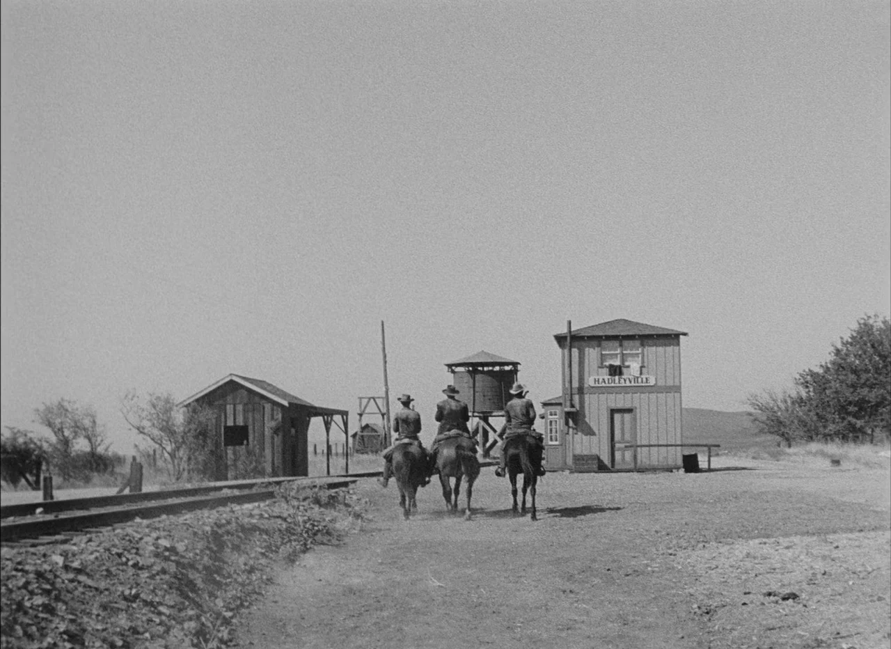High Noon (1952) movie still: wide — A desolate 19th-century frontier depot with a wooden water tower and station building. Three mounted cowboys…