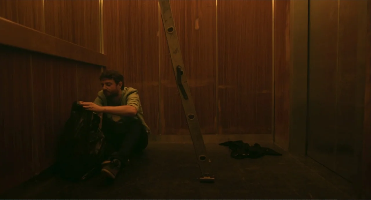 The Lift (2021) movie still: wide — An elevator interior features wood-paneled walls and a metal ladder leaning against one side. A man sits on the…