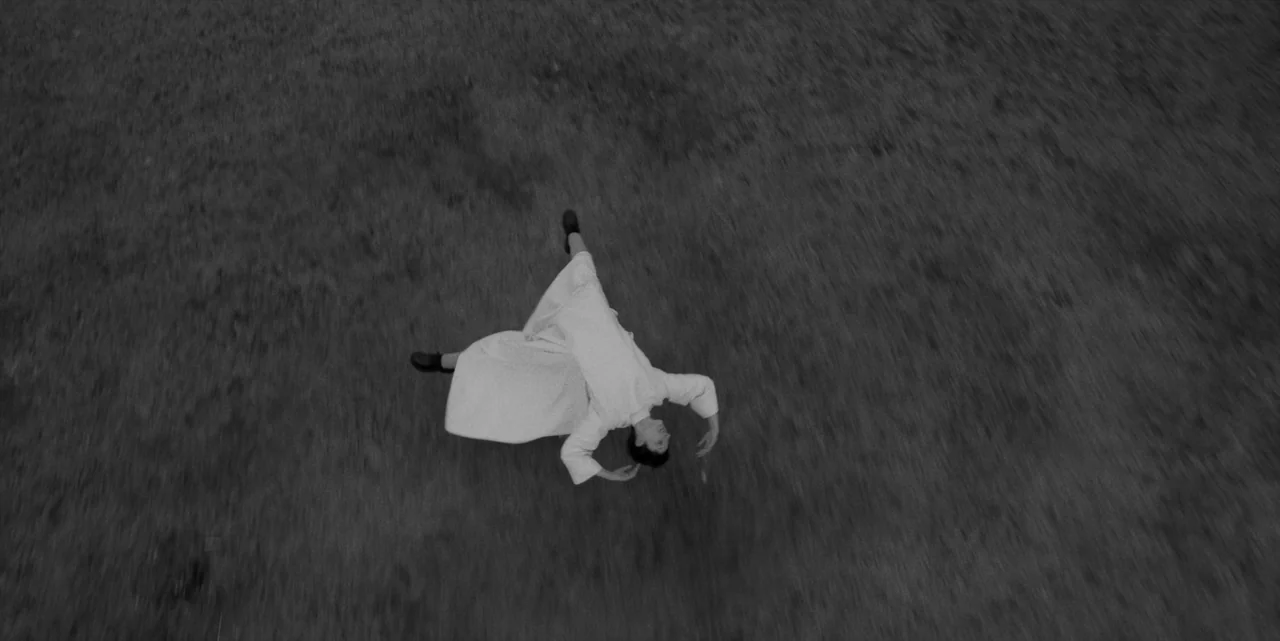 El Conde (2023) movie still: wide — An open grassy field stretches out under an overhead perspective. A person in a white robe lies flat on the…