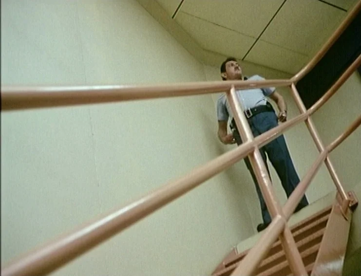 Ghosts… Of The Civil Dead (1988) movie still: fullbody — A utilitarian stairwell with plain white walls and pale metallic railings. A man in a…