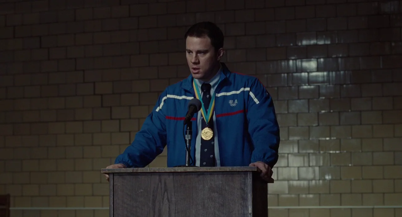 Foxcatcher (2014) movie still: medium — A dimly lit room features a tan brick wall as the background. A man stands at a podium wearing an Olympic…