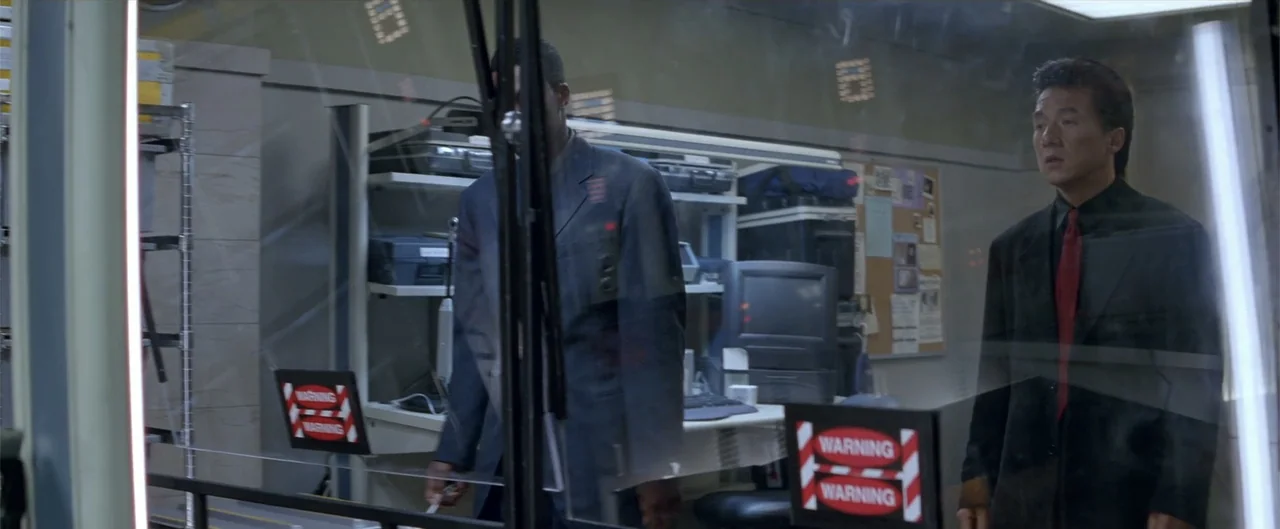 Rush Hour (1998) movie still: medium — A sterile laboratory room contains computer equipment, storage shelves, and warning signs on glass. Two men…