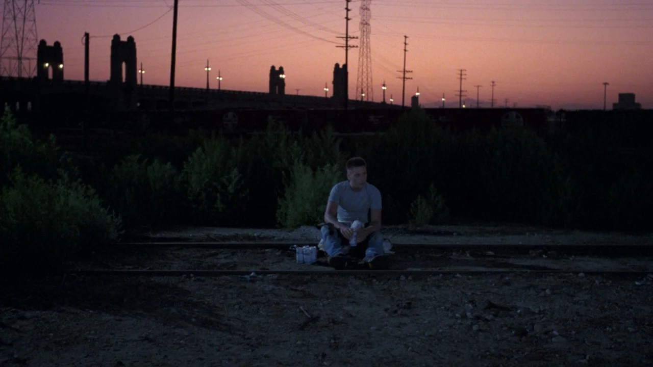 Repo Man (1984) movie still: wide — A man sits alone on abandoned railroad tracks during a dim sunset. Industrial bridge structures and power lines…