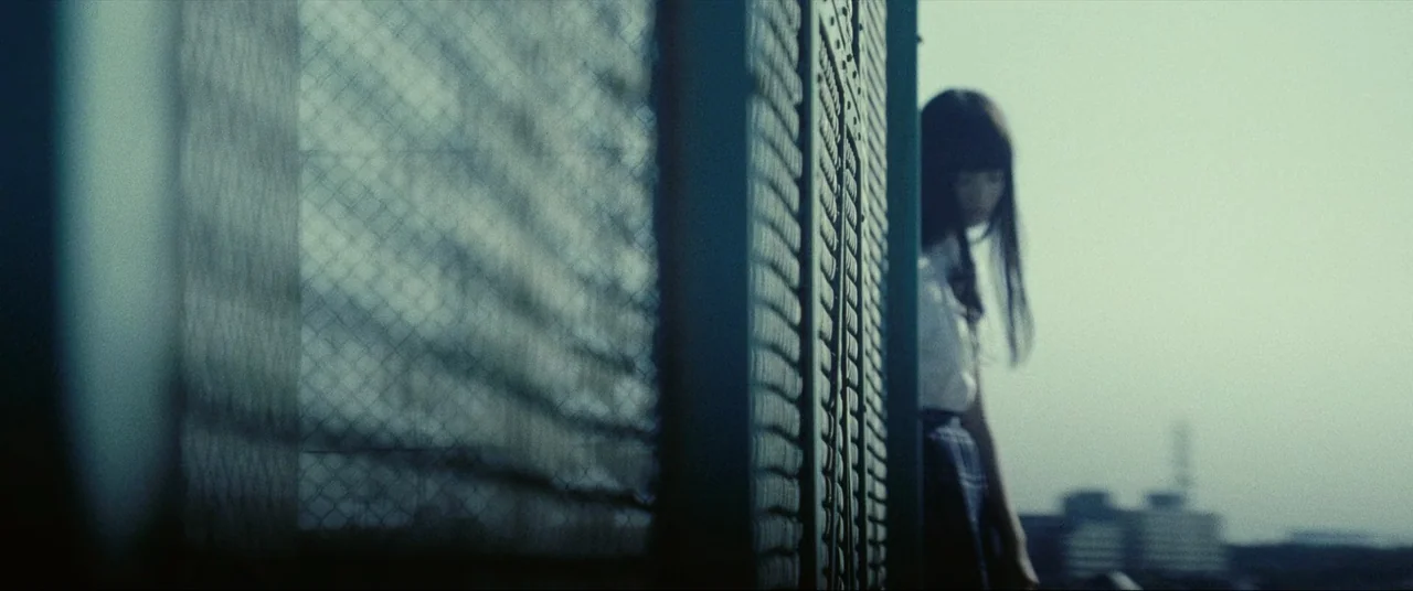 The World of Kanako (2014) movie still: medium — An outdoor area features a prominent chain link fence in the foreground. A teenage girl in a school…