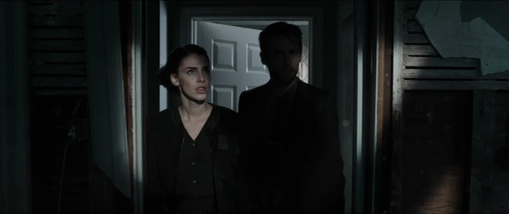 Abattoir (2016) movie still: medium — A dark, dilapidated hallway with exposed wall studs and unfinished plaster surfaces. A man and a woman stand…