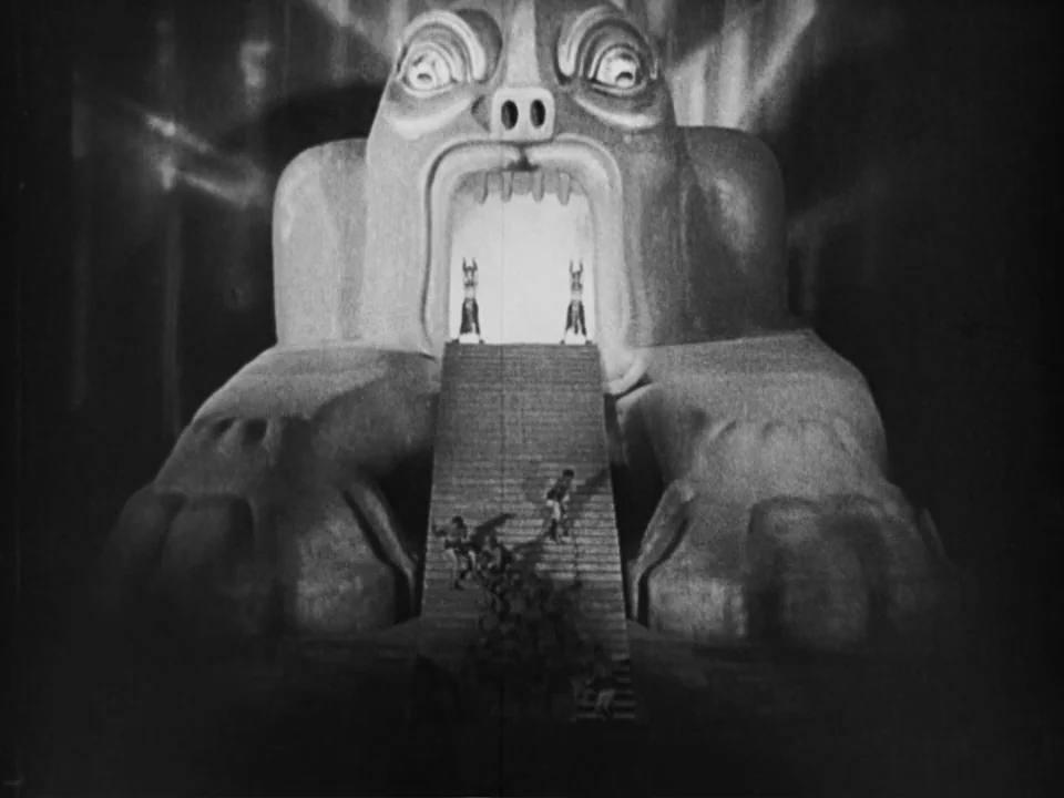 Metropolis (1927) movie still: wide — A colossal stone statue features an open mouth serving as the entrance to a temple. Small figures ascend the…