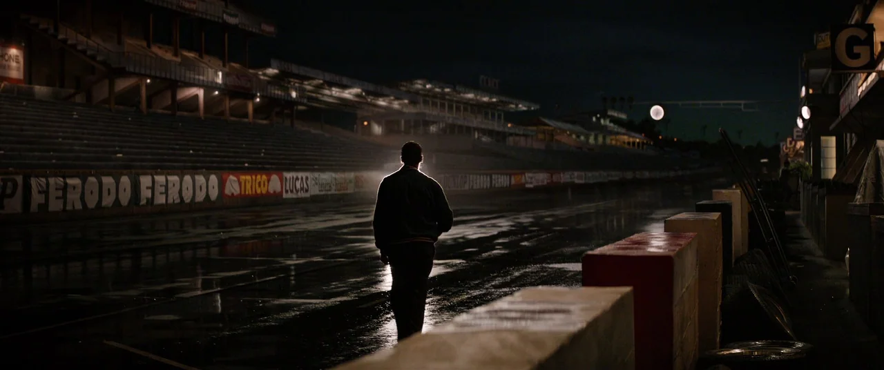 Ford V Ferrari (2019) movie still: wide — A dark, wet racetrack at night features long rows of empty grandstands and barriers. A solitary figure…