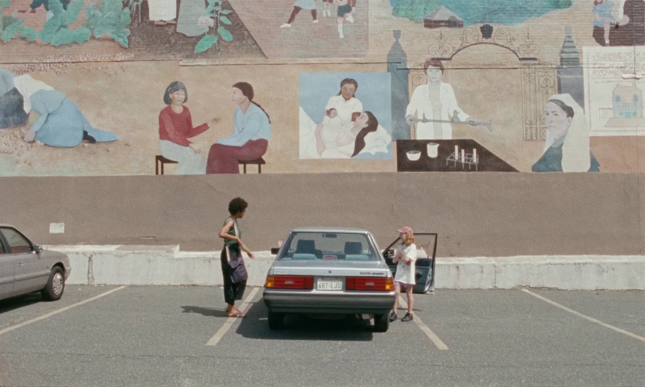 Janet Planet (2023) movie still: wide — An asphalt parking lot sits before a large painted brick mural featuring historical scenes. A woman and a…