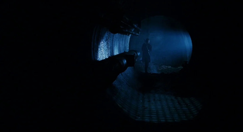 Mimic (1997) movie still: wide — A dark subterranean concrete tunnel features a large cylindrical opening at the end. A lone figure stands in the…