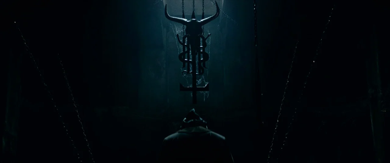 The Divine Fury (2019) movie still: wide — A dark stone vault features cobwebs and hanging metal chains in the shadows. A hooded figure stands…