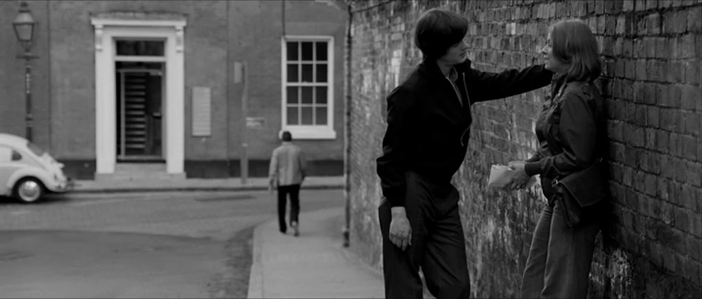 Control movie still: fullbody — A man and woman stand on a sidewalk next to a textured brick wall. A third person walks away in the distance on a…