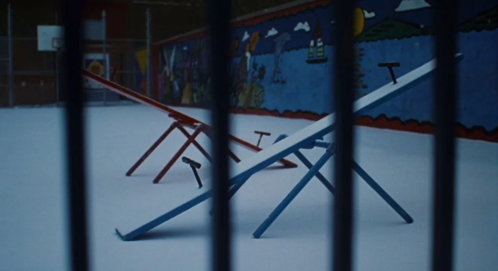 Mimic (1997) movie still: wide — A snowy playground at night is enclosed by dark vertical metal bars in the foreground. Two empty seesaws stand on…
