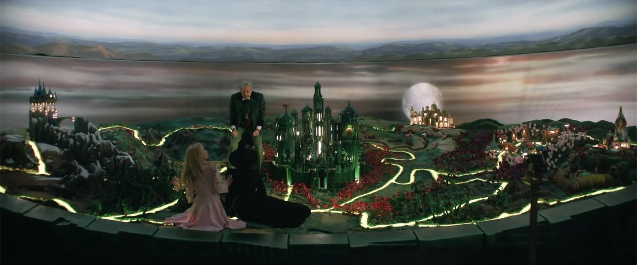 Wicked (1998) movie still: wide — A dimly lit room houses an elaborate illuminated diorama of a fantasy kingdom. Three people stand and sit around…