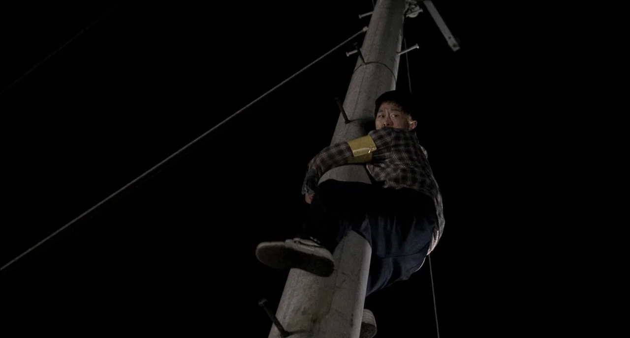Memories of Murder (2003) movie still: medium — A concrete utility pole extends upward into the pitch black night sky. A man climbs the pole using…