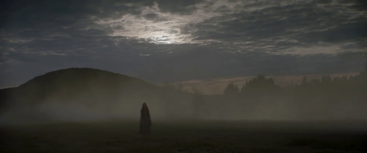 Ophelia (2018) movie still: wide — A vast, open field is shrouded in thick, low-lying ground mist beneath an overcast sky. A lone figure in a dark,…