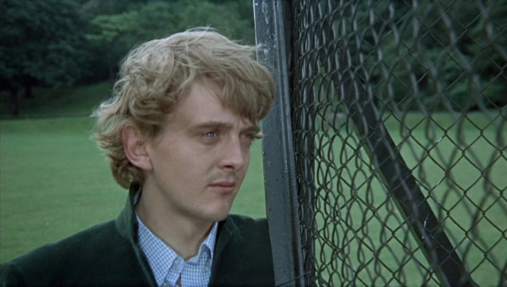 Blow-Up (1966) movie still: medium — A man stands next to a chain-link fence in a grassy public park area. He leans his head sideways against the…