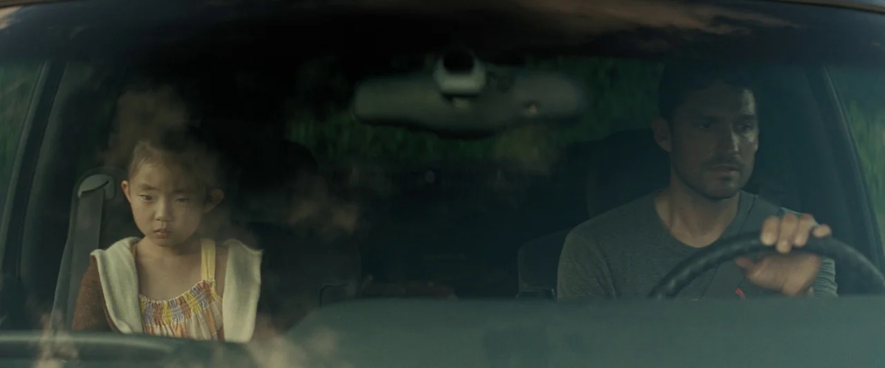 Knock at the Cabin (2023) movie still: medium — The interior of a car shows a man behind the wheel and a young girl. They both look forward while…