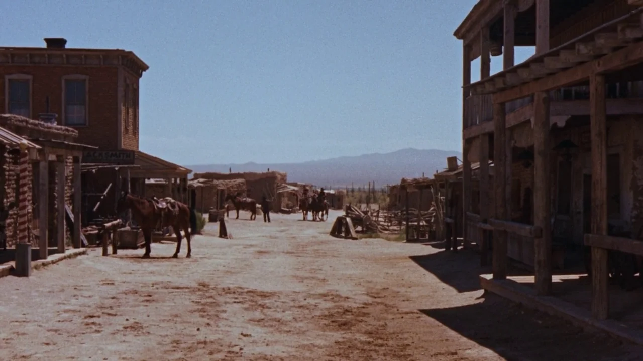 Rio Bravo (1959) movie still: establishing — A dusty dirt street in a western town bordered by wooden and adobe buildings. A few horses are tied to…