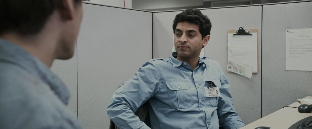 Office Uprising (2018) movie still: medium — A standard office cubicle contains grey fabric walls and a desk surface. A man in a blue shirt sits in…
