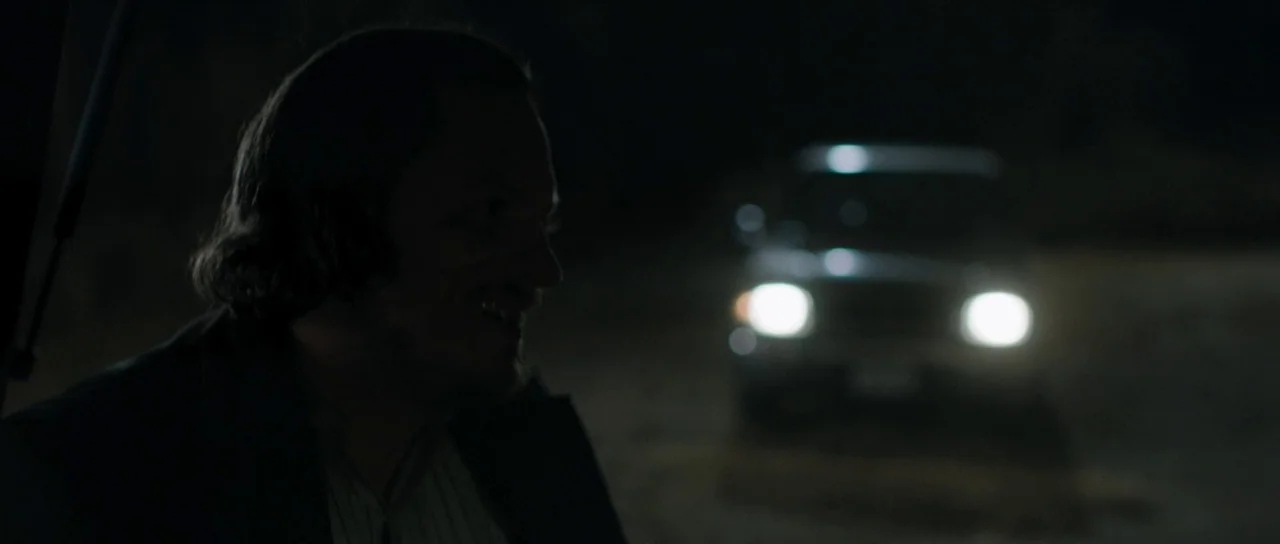 Joe (2013) movie still: medium — A dark outdoor setting at night with a person standing in the shadows. A car is visible in the distance with its…