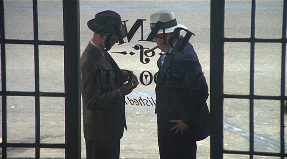 Thieves Like Us (1974) movie still: medium — Two men in vintage suits stand inside a bank entrance behind a glass door. They face each other while…
