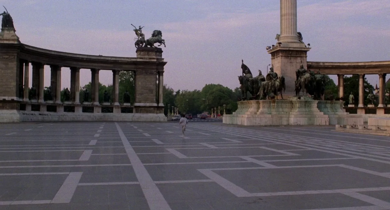 Etoile (1989) movie still: wide — A wide, open paved plaza features a large stone monument with bronze statues. A solitary person jogs across the…