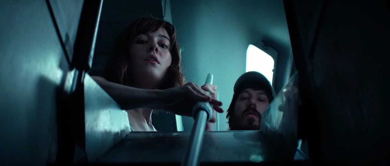 10 Cloverfield Lane (2016) movie still: medium — A cramped, dimly lit interior space with metallic walls and a rectangular hatch opening. Two people…