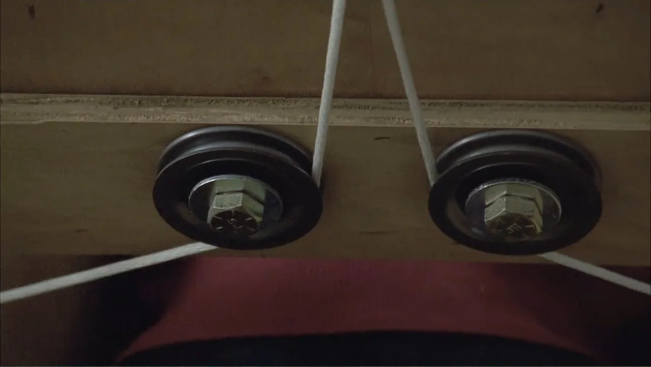 Temple Grandin (2010) movie still: closeup — A wooden board features two metal pulleys with hex bolts attached to it. A white cord threads through…