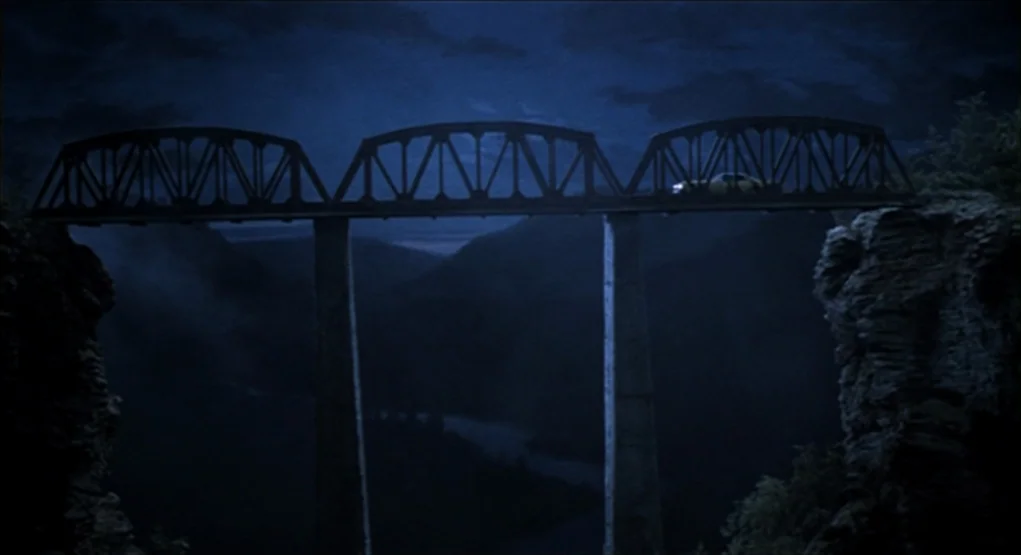 Evil Dead II (1987) movie still: establishing — A metal truss bridge spans a deep, dark ravine at night under a cloudy sky. A single yellow vehicle…