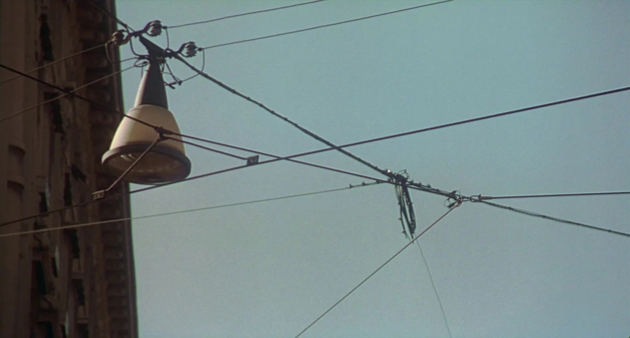 Making of: I Am Love (2009) movie still: establishing — An overhead view of trolley wires and a street lamp against a pale sky. The wires crisscross…