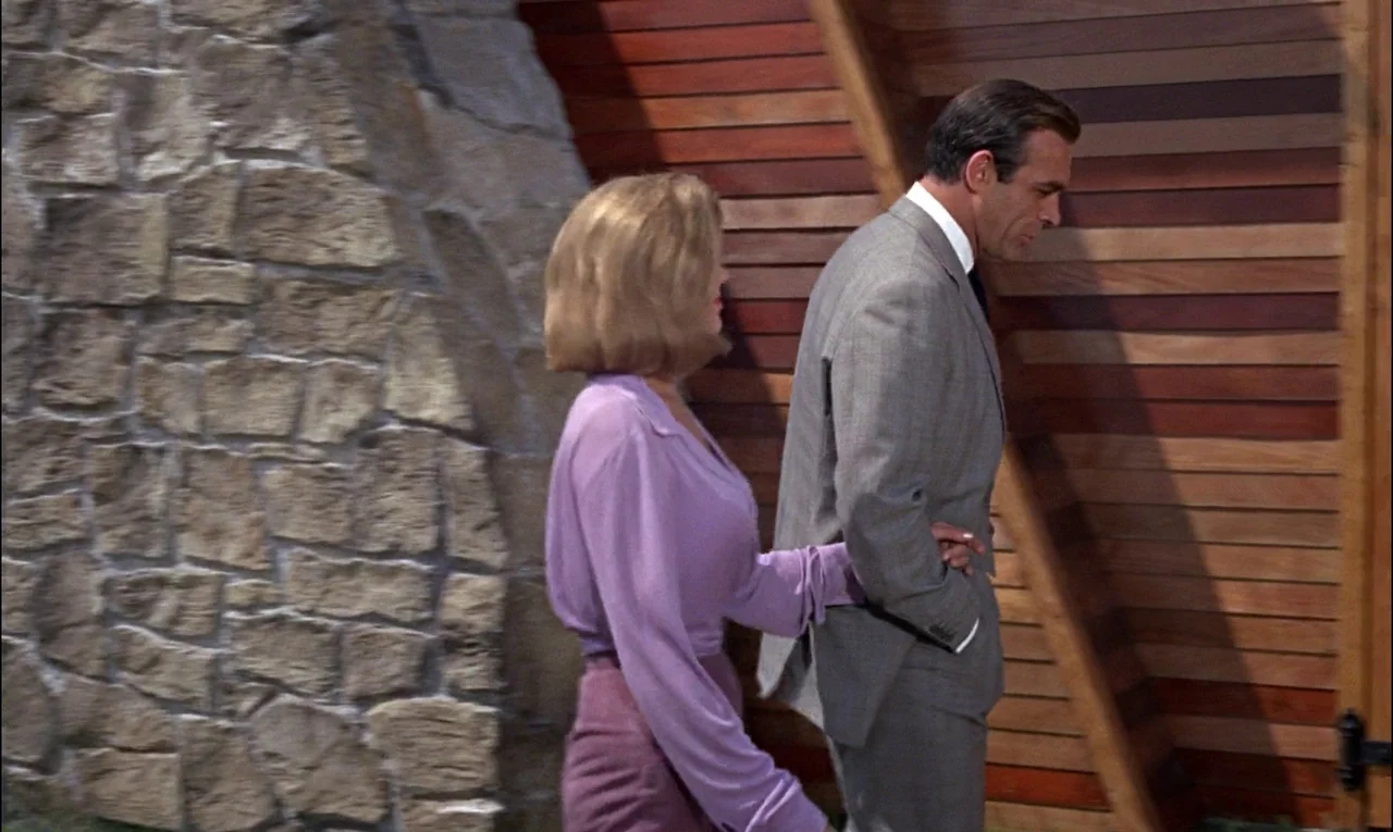 Goldfinger (1964) movie still: medium — A modern room features a textured fieldstone wall and horizontal wooden paneling. A blonde woman in a purple…