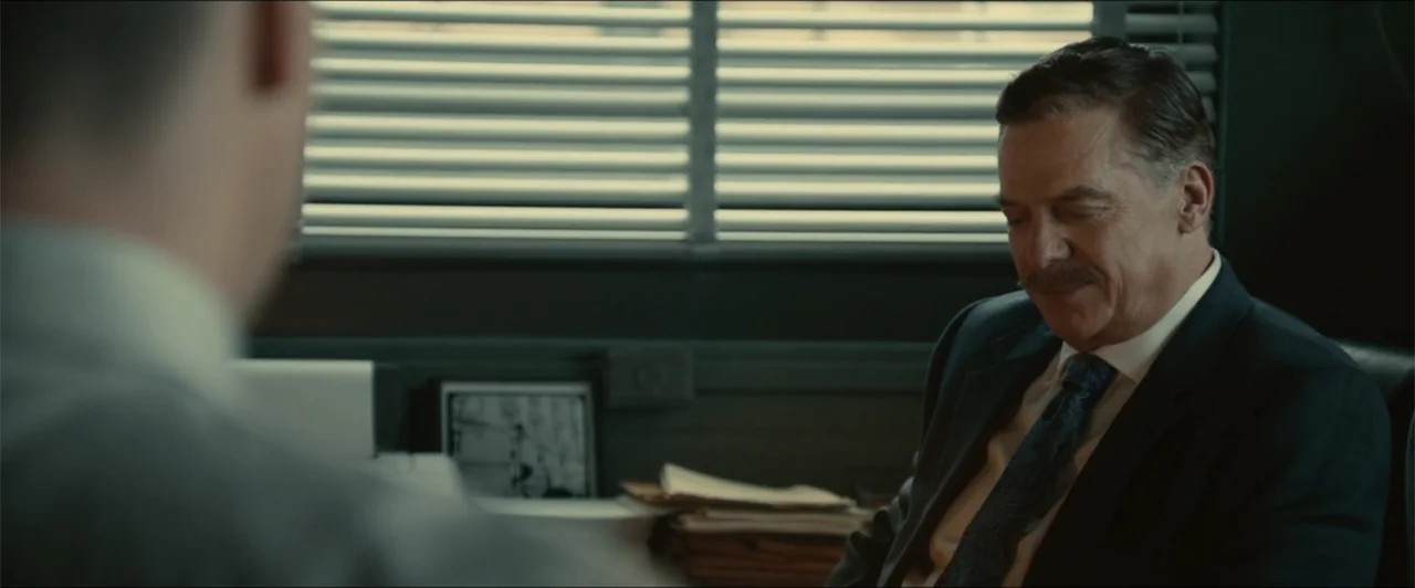 Exposed (2016) movie still: medium — A dark professional office with horizontal blinds covering a window behind the desk. A middle-aged man in a…