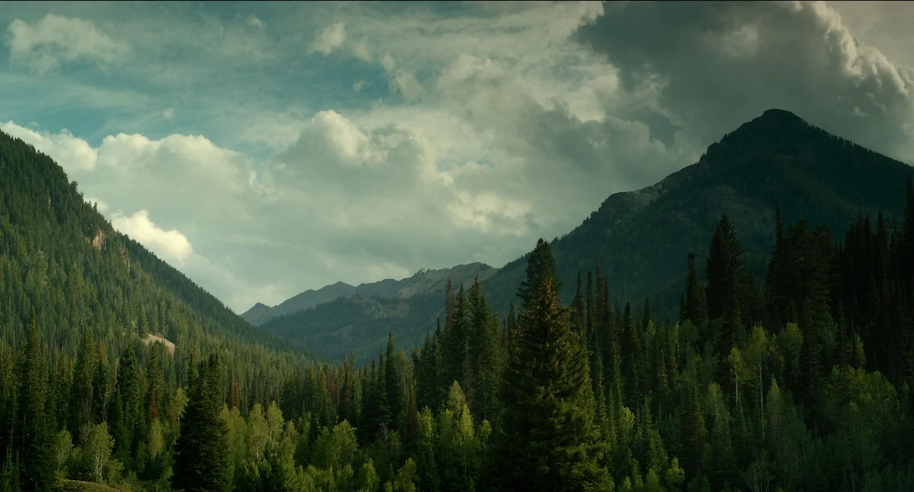 Trim Season (2023) movie still: establishing — A deep valley is nestled between dense pine-covered mountains under a cloudy sky. The landscape…