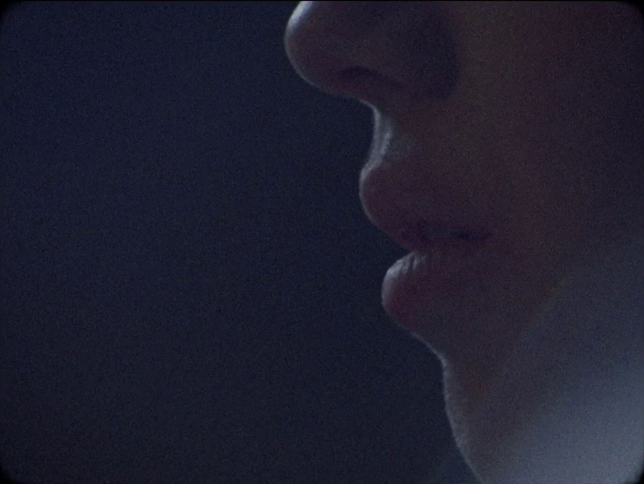 The Devil’s Doorway (2018) movie still: closeup — The frame shows a close-up of a human profile in a very dark setting. The nose, lips, and chin of…