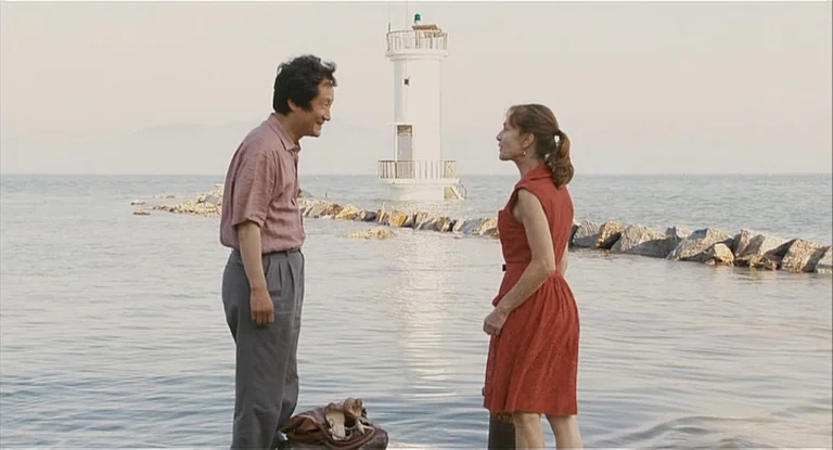 In Another Country (2012) movie still: fullbody — Two people stand on a narrow stone path surrounded by water. A small white lighthouse stands in…