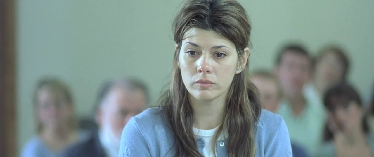 In the Bedroom (2001) movie still: medium — A sterile courtroom with blurred figures seated in rows behind a woman. The woman sits in the foreground…