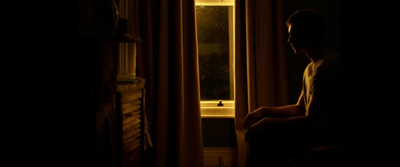 The Cured (2017) movie still: medium — A dark room features a window with heavy curtains pulled to the sides. A young man sits in profile while…