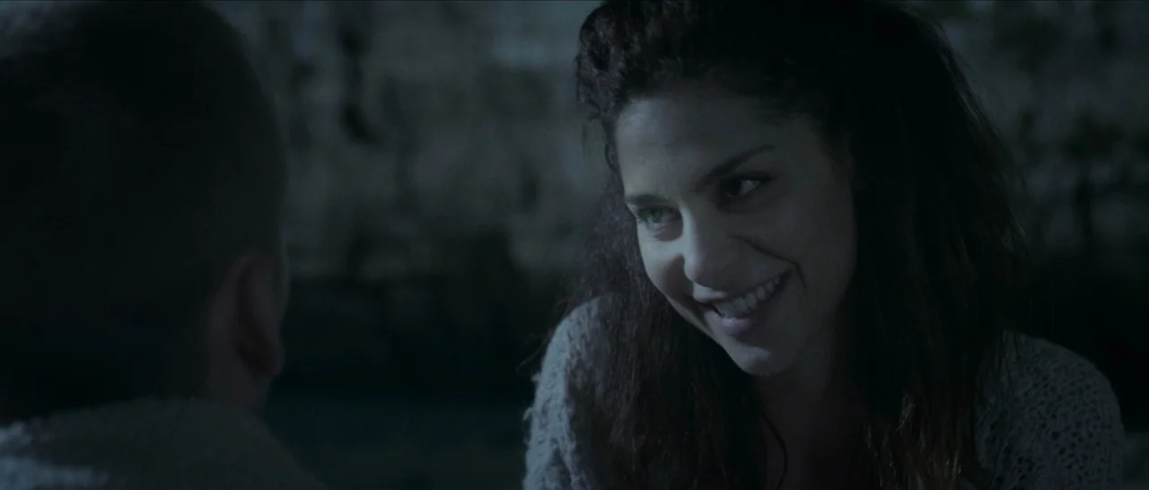 Spring (2014) movie still: medium — A dark outdoor space features a woman smiling at an unseen man in the foreground. The background consists of a…