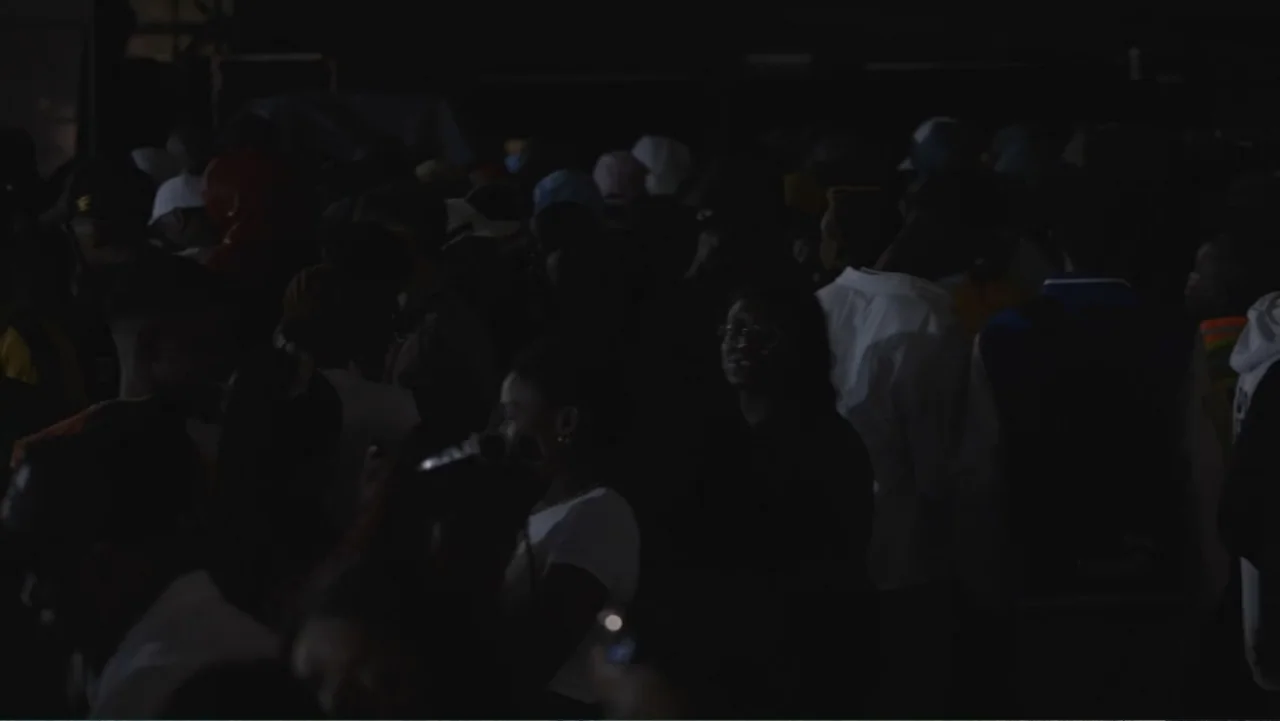 African Throne World Tour (2024) movie still: wide — A dense crowd of people stands in a dark, outdoor urban environment at night. The individuals…